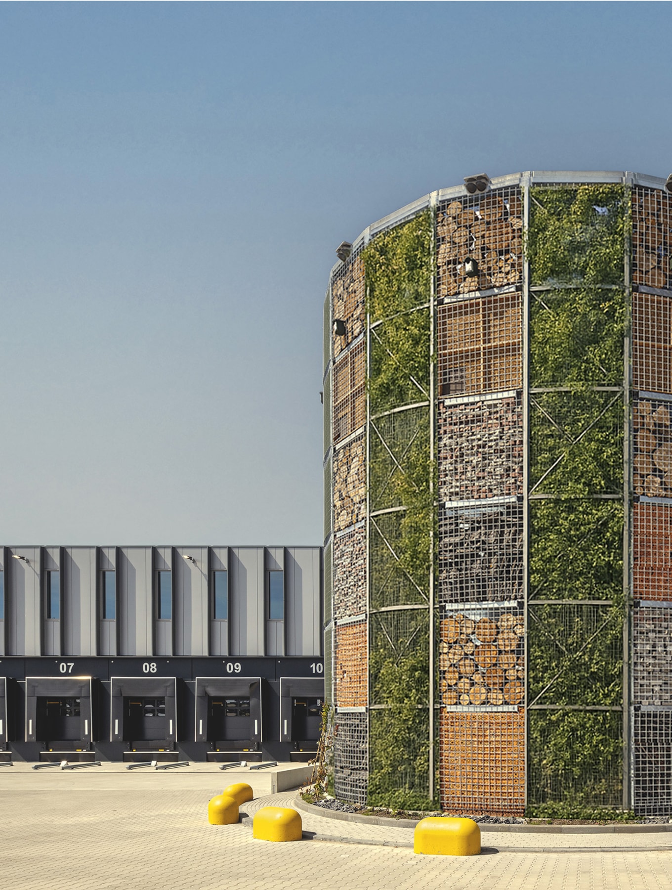 A cylindrical structure with stacked natural materials and greenery stands near a modern industrial building with loading bays.