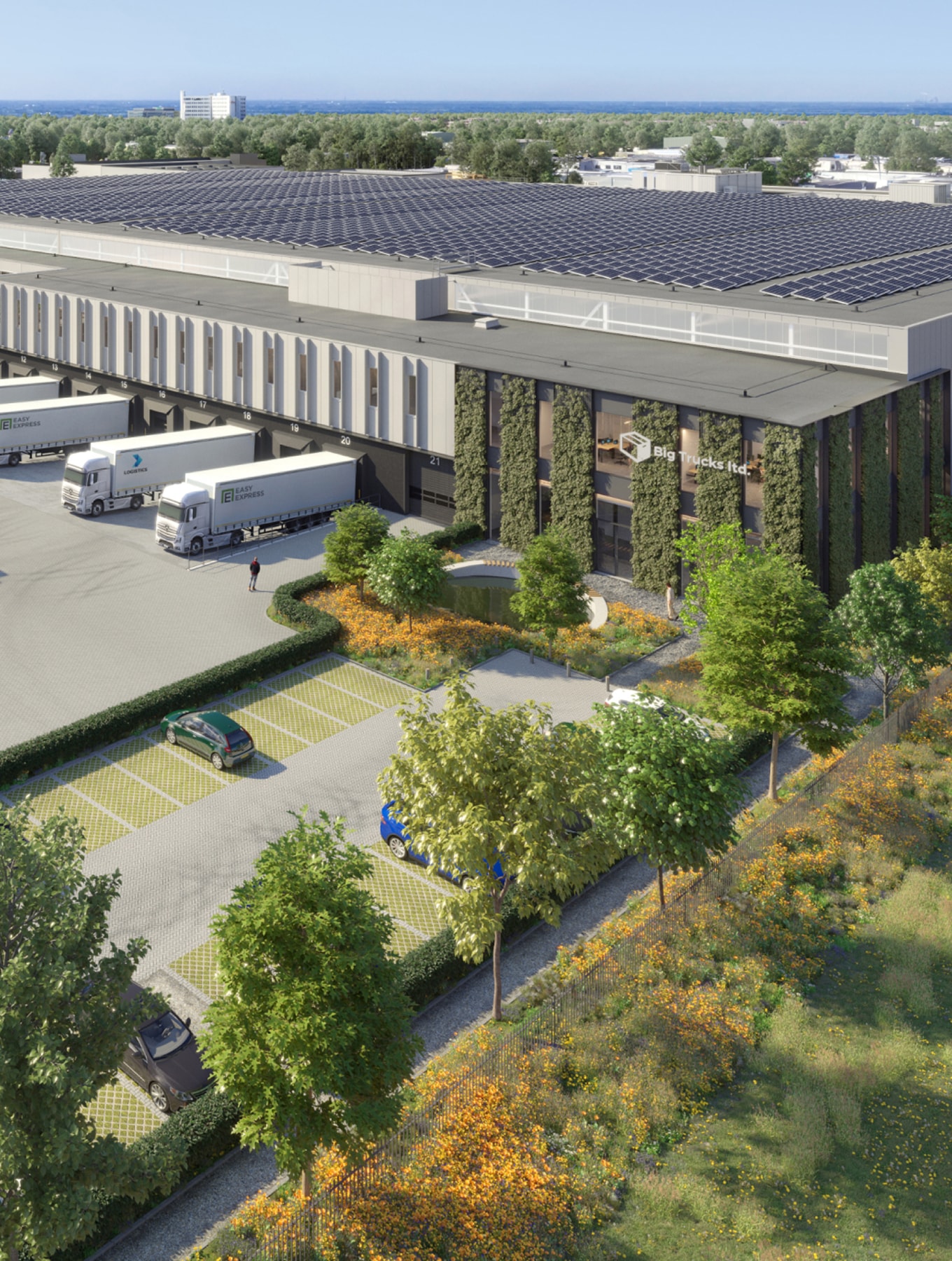 Aerial view of a modern warehouse with solar panels, trucks parked outside, surrounded by greenery and trees, under a clear blue sky.