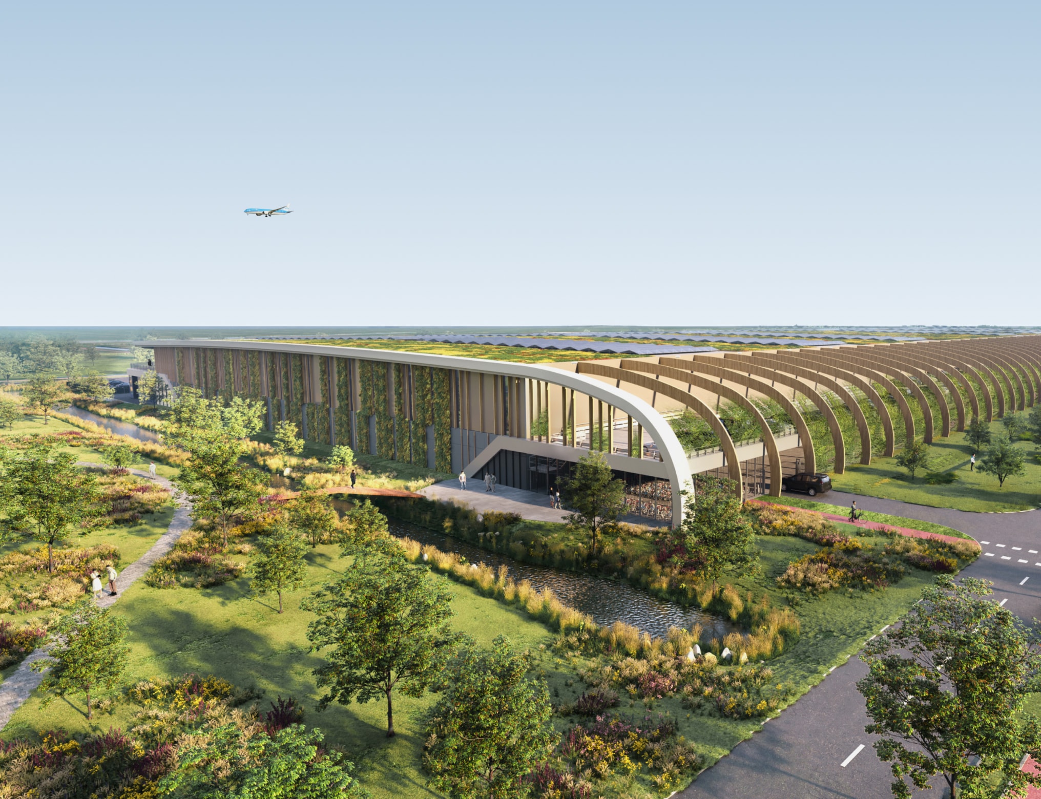 Modern warehouse with a curved, green roof design, surrounded by trees and gardens. An airplane is flying overhead in a clear sky.